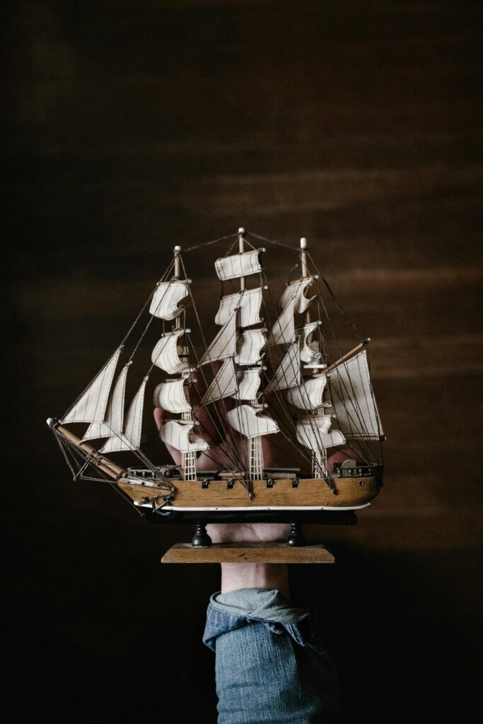 A hand in denim holds a vintage wooden model ship against a dark background.