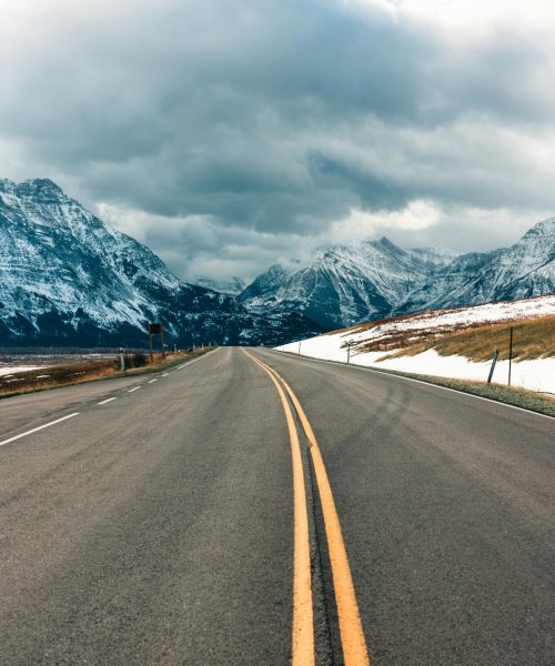 Open road leading to snow-covered mountains in Waterton Park, Alberta, Canada under cloudy skies.