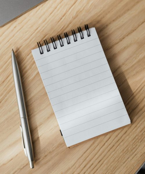From above of small opened notepad and stylish silver pen placed on wooden table near laptop